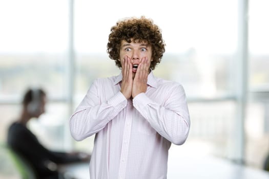 Portrait of scared shocked young man with curly hair. Office setting in the background.