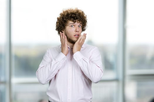 Portrait of surprised young guy with curly hair touching his face. Indoor window in the background.