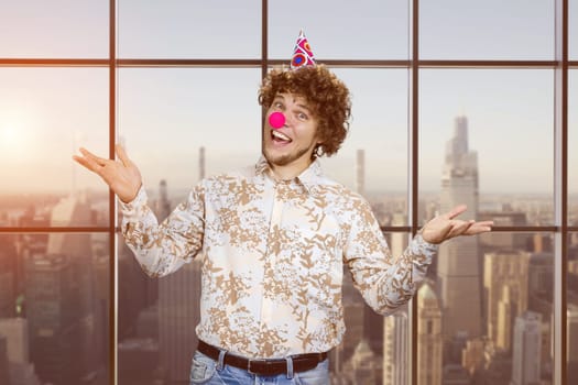 Portrait of a happy young man with clown nose. Indoor window with evening sunset view in the background.