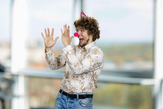 Happy young caucasian guy with curly hair wearing a clowns nose. Indoor window in the background.