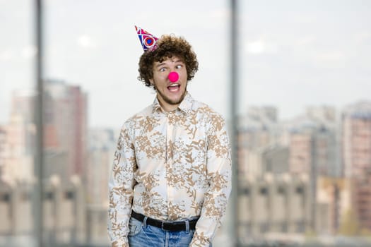 Happy young caucasian guy with clown nose fooling around making funny faces. Indoor window in the background.