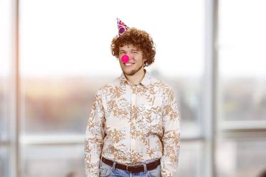 Portrait of happy young guy with clown nose. Indoor window in the background.