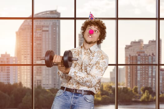 Portrait of a young clown fooling around with dumbbell indoors. Checkered window in the background.