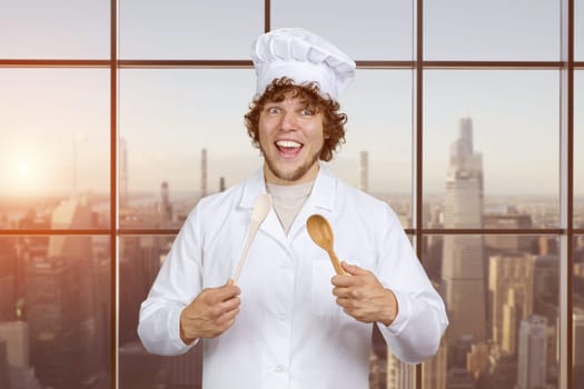 Portrait of a happy young caucasian male cook with a wooden spoon. Checkered window with cityscape in the background.