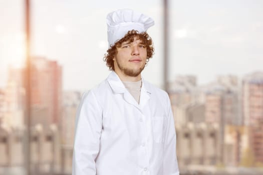 Portrait of a young male cook in white uniform standing indoors. Window with cityscape view in the background.