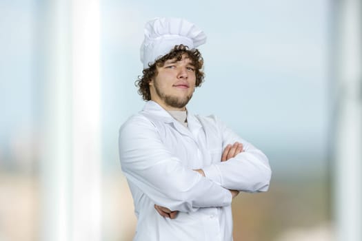 Portrait of a young male cook with folded arms. Indoor window in the background.