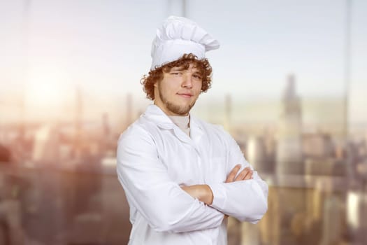 Portrait of a young male cook in white uniform standing indoors with folded arms. Window with cityscape view in the background.