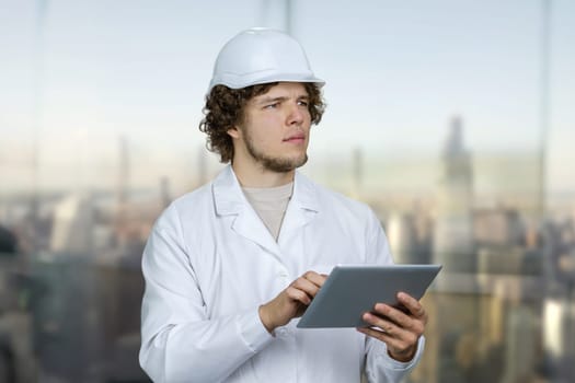 Portrait of a young man in white uniform and white helmet holding tablet pc device. Blurred cityscape background.