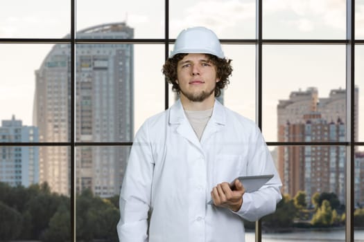 Portrait of a young construction worker in white uniform holding tablet pc device. Checkered window with cityscape view in the background.