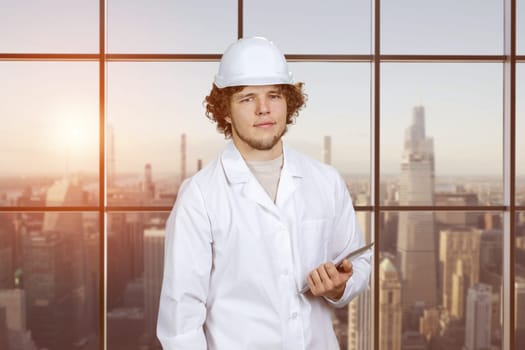 Portrait of a young guy in white uniform holding tablet pc device. Indoor window with cityscape view in the background.