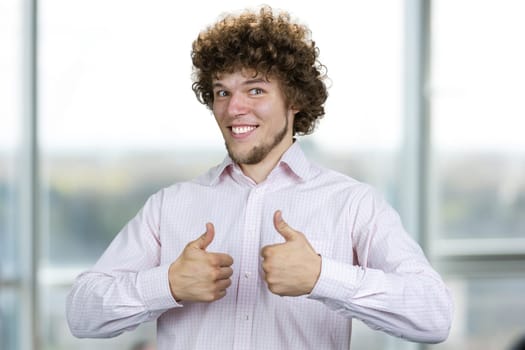 Happy smiling young man with curly hair shows both thumbs up. Indoor window in the background.