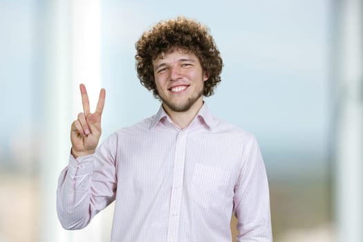 Portrait of a happy young man with curly hair shows victory gesture sign. Indoor window in the background.