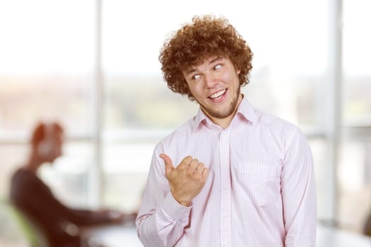 Portrait of happy smiling young man with curly hair pointing back with his thumb. Office environment in the background.