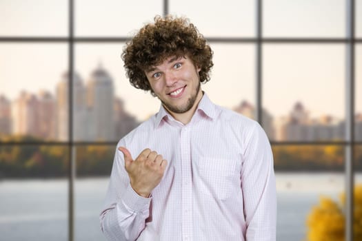 Portrait of happy cheerful young man with curly hair pointing back with his thumb. City river in the background.
