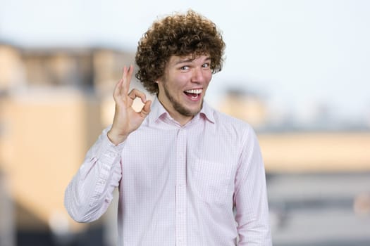 Portrait of a happy young man with curly hair shows okay gesture sign. Blurred urban background.