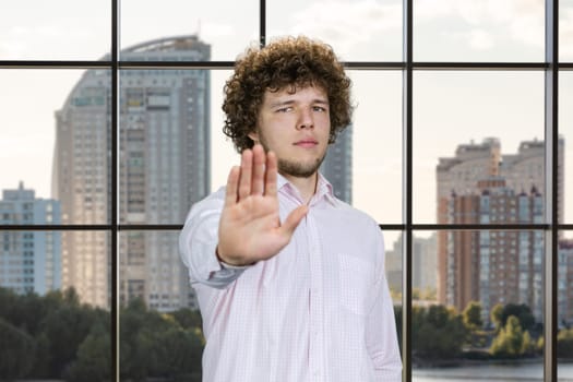 Portrait of a young man with curly hair shows his stop gesture sign. Checkered window in the background.