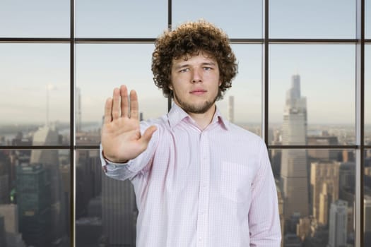 Serious young man with curly hair shows his stop gesture sign. Checkered window with cityscape view in the background.