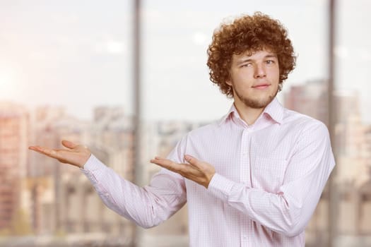 Portrait of a young man with curly hair advertising copy space with both hands. Cityscape in the background.