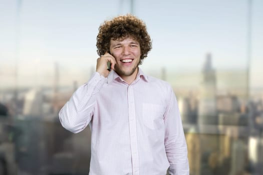 Portrait of a young caucasian guy in white shirt talking on the phone. Indoor window with blurred cityscape view in the background.