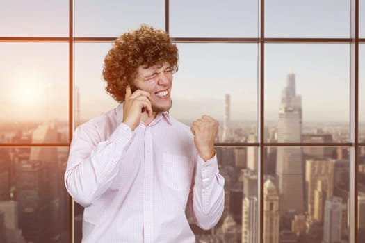 Portrait of a young happy caucasian man talking on the phone rejoicing success. Checkered window with evening cityscape view.