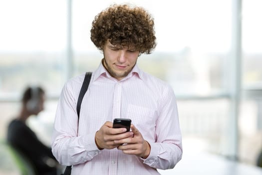 Portrait of a young man with curly hair using his smartphone in the office. Indoor window in the background.