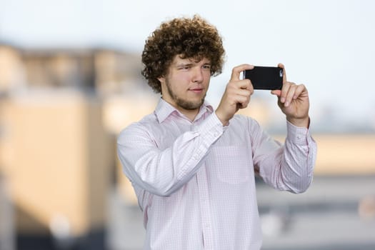 Portrait of a young tourist with curly hair making a photo on his smartphone camera. Blurred urban scape in the background.