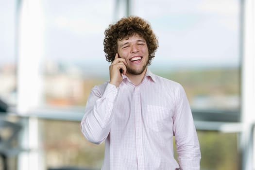 Portrait of a young man in white shirt talking on the phone and laughing. Indoor window in the background.