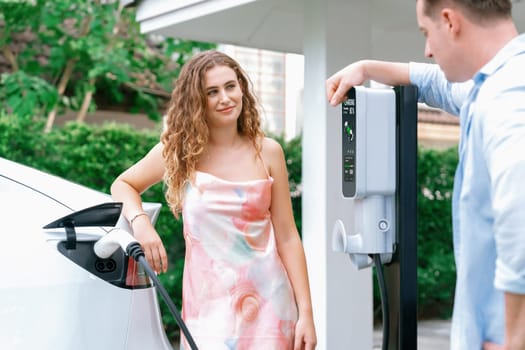 Happy and lovely couple with eco-friendly conscious recharging electric vehicle from EV home charging station. EV car technology utilized for residential home to future sustainability. Synchronos