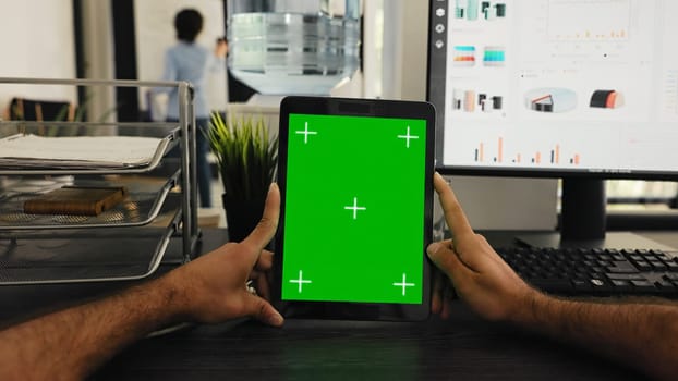 POV of businessman holding tablet with greenscreen template, looking at chromakey display and sitting coworking space desk. Person examining isolated mockup copyspace on gadget screen.