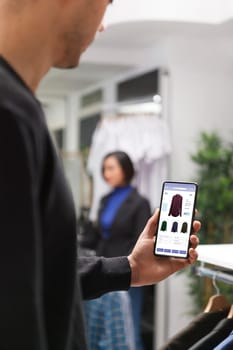 Clothing store customer male hand grasping smartphone and checking apparel on boutique website. Caucasian client using mobile device for browsing clothing items in a shopping mall.