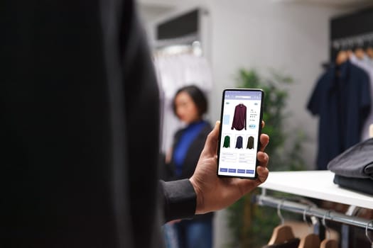 Close-up shot of a person browsing through clothing items on a smartphone screen, emphasizing the role of technology in shopping experience. Photo focus on mobile device displaying fashion collection.