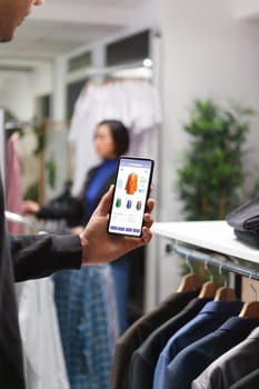 Smartphone being held by caucasian person in shopping mall. Close-up of mobile device displaying clothing virtual store grasped by white male customer in a modern boutique.