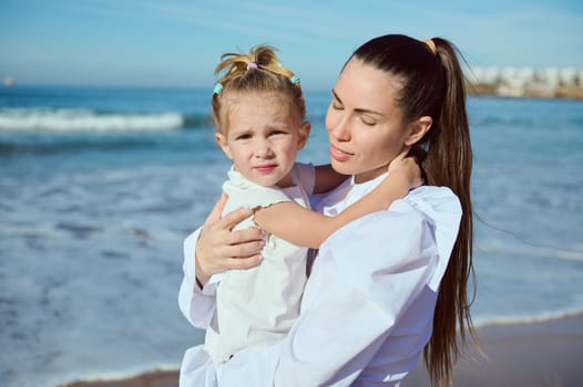 Authentic portrait of a beautiful young Caucasian woman carrying her adorable little child girl, enjoying happy family pastime together while walking on the beach. People. Lifestyle. Leisure activity