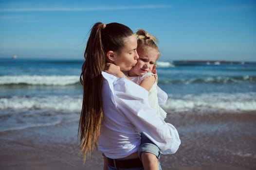 Authentic portrait of a beautiful young Caucasian woman carrying, kissing and hugging her little daughter, enjoying happy time together while walking on the beach. People. Lifestyle. Leisure activity