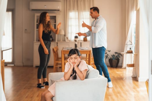 Annoyed and unhappy young girl sitting on sofa trapped in middle of tension by her parent argument in living room. Unhealthy domestic lifestyle and traumatic childhood develop to depression.Synchronos