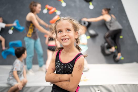 Smiling little girl climber standing arms crossed in sports center. Female child looking up during rock climbing class. Essence of pursuing hobbies.