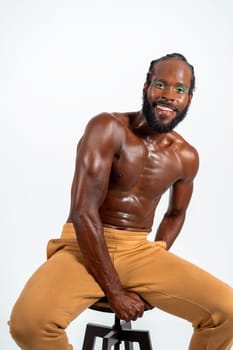 Portrait of shirtless gay man with eyeshadow sitting on stool. Confident muscular bearded man smiling on white background. Happy African American gay person with bright makeup.