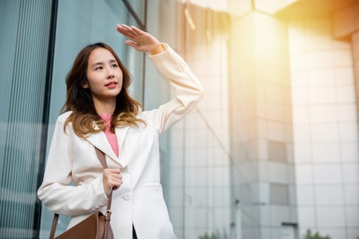 Portrait of confidence young businesswoman standing outside office building in city raise her hand to shade sun UV. Happy woman wearing white suit jacket with brown bag at sunlight outdoors. sun block