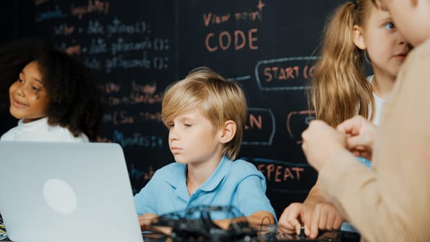 Closeup of boy using laptop programing engineering code and writing program while group of smart diverse student standing surrounded by friend in STEM technology classroom at blackboard. Erudition.