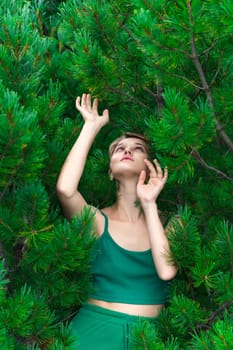 Nosy European woman pulls both hands up and looking up, poses against background evergreen shrub of Stone Pine. Sensual Caucasian adult female with short hair dressed in green crop top. Part of series