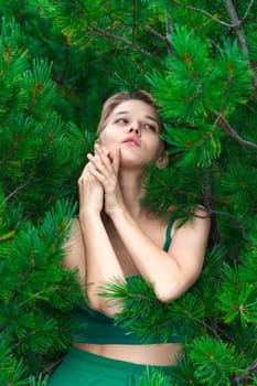 Shy attractive blonde woman poses against background of an evergreen shrub of Japanese Stone Pine. Elegant European model dressed in green cropped top, looking away. Part of series