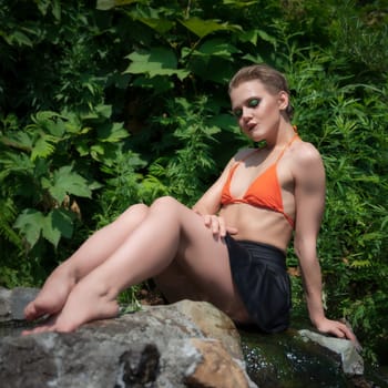 Woman relaxing sitting legs stretched sensually on rocky edge of outdoor pool with geothermal water. Adult female with makeup closed eyes, bit lower lip, dressed in orange bikini top, black mini skirt