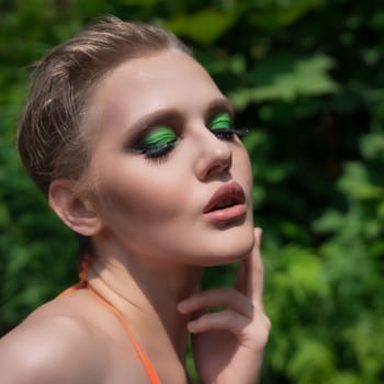 Close-up portrait of blonde woman with closed eyes and bright makeup posing against background of green leaves in summer forest. Fashionable adult female Caucasian ethnicity has sensually parted mouth