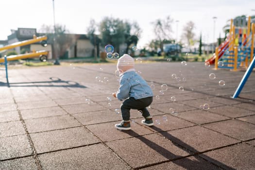 Little girl squats down on the playground and catches soap bubbles. Back view. High quality photo