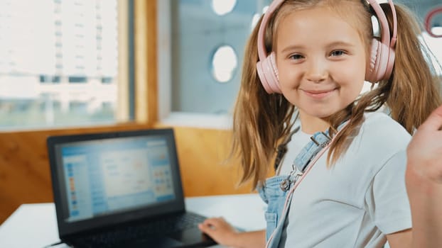 Smiling girl looking while waving hand at camera with laptop placed on table. Child wearing headphone smiling while laptop screen show system programing or coding program in STEM class. Erudition.