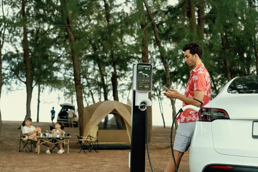 Outdoor adventure and family vacation camping in nature travel by eco friendly car for sustainable future. Lovely sit on trunk, charging EV car with EV charging station in campsite. Perpetual