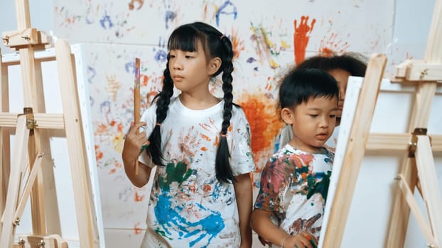 Playful student painted or draw canvas at stained wall with diverse friends in art lesson. Group of happy multicultural children working or create artwork at messy room. Creative activity. Erudition.