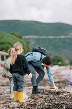 Little girls with bags look at their mother collecting trash on the beach. Back view. High quality photo