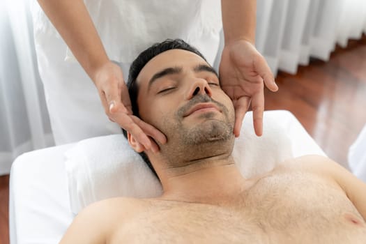 Caucasian man enjoying relaxing anti-stress head massage and pampering facial beauty skin recreation leisure in dayspa modern light ambient at luxury resort or hotel spa salon. Quiescent