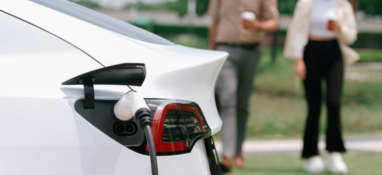 Young couple with coffee cup, recharge electric car battery from EV charging station. Sustainable and eco friendly EV car with urban and shopping lifestyle. Panorama Expedient